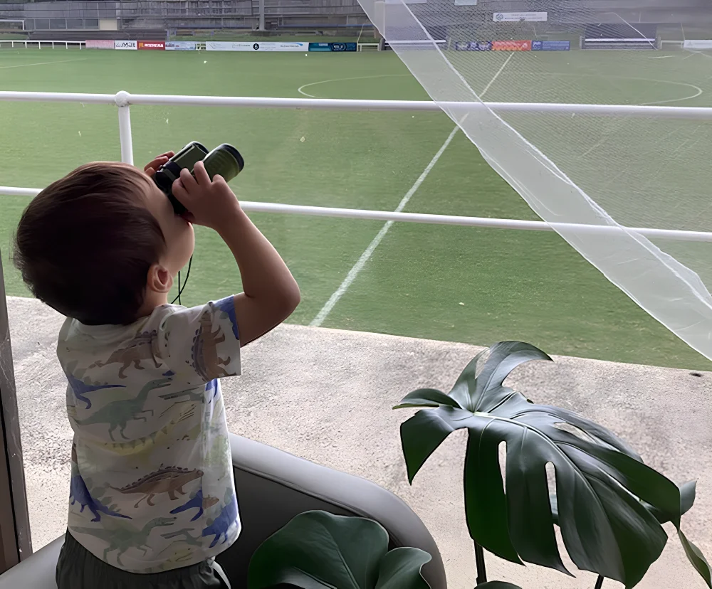 Child standing by a railing looking through binoculars at a sports field outdoors children's centre Woonona