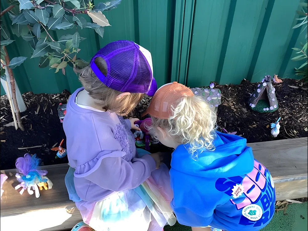 Two children sitting beside a garden bed playing with small fairy figurines and toys outdoors at children's centre Woonona