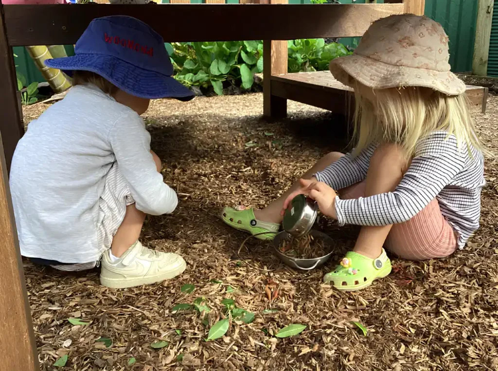 Two children sitting under a wooden bench playing with woodchips and a metal bowl in an outdoor play area at children's centre Woonona