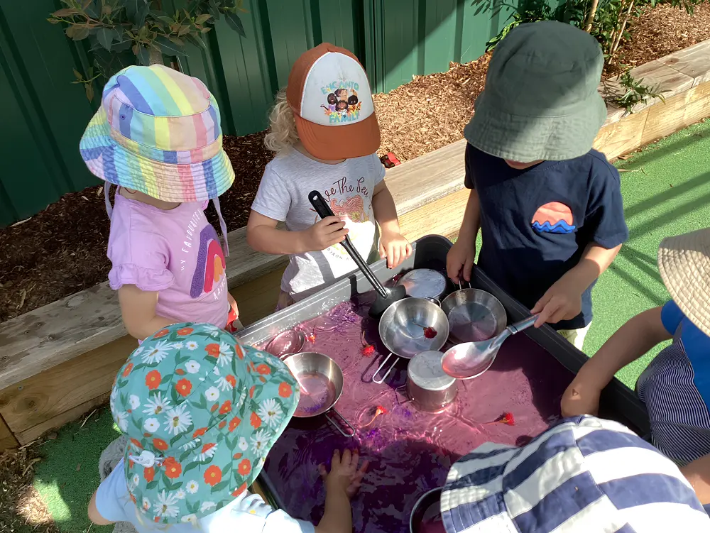 Children wearing sun hats enjoying sensory water play with bowls and utensils at an outdoor preschool table.