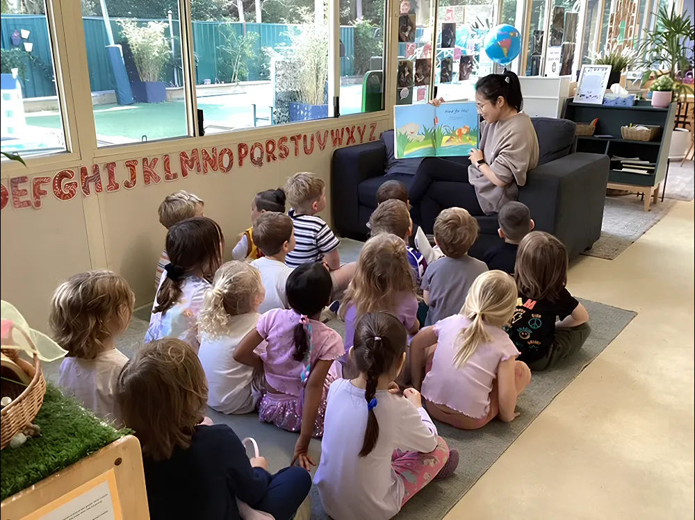 Teacher sitting in a classroom reading a picture book to a group of preschool children seated on the floor at children's centre Woonona