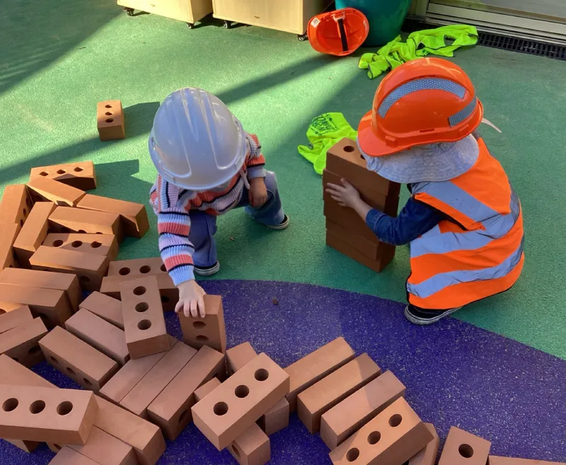 Children wearing construction helmets and high visibility vests building a tower with large toy bricks during imaginative construction play.